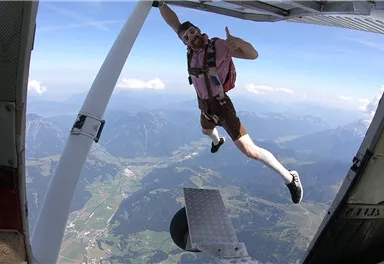 Ein mutiger Mensch springt aus einem Flugzeug und macht einen Daumen hoch. Die Aussicht auf die Berge und das grüne Tal ist atemberaubend.