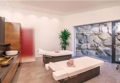 Ein modernes Wellnesszimmer mit zwei Massagetischen und einem großen Fenster, das auf eine Steinwand blickt. Die Einrichtung ist minimalistisch, mit Pflanzen und eleganten Holzelementen.
