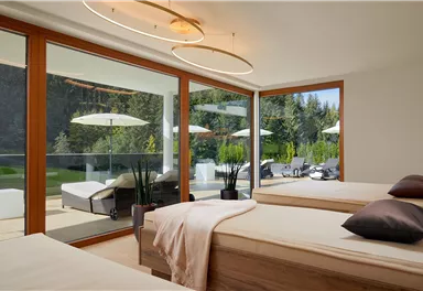 A modern bedroom with two beds and large windows. Outside, lounging chairs and sun umbrellas are visible, surrounded by nature.