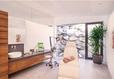 Ein moderner Behandlungsraum mit einer Liege und einem großen Fenster, das auf einen Steinwandblick zeigt. Der Raum ist hell und minimalistisch eingerichtet, mit Pflanzen und einigen kosmetischen Produkten.