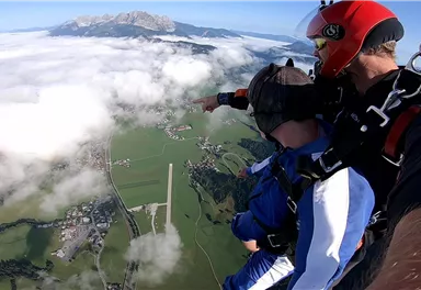 Ein Tandemsprung aus dem Flugzeug über einer grünen Landschaft und Wolken. Im Hintergrund sind die Berge zu sehen.