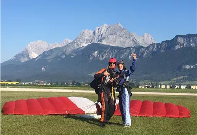 Zwei Paragleiter stehen lächelnd auf einer Wiese mit einem roten Fallschirm im Hintergrund. Im Hintergrund sind majestätische Berge und ein klarer blauer Himmel sichtbar.