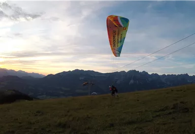 A paraglider glides over a mountain landscape at sunset. The sky is colorful and the mountains are visible in the background.
