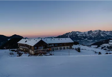 A rustic mountain cabin in the snow in front of majestic mountains. The sky displays soft colors at sunset.
