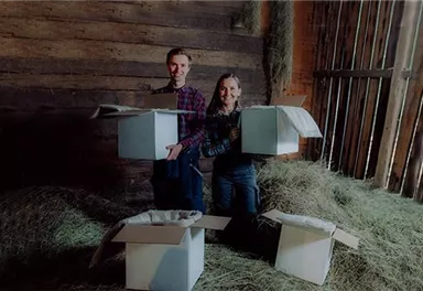 Zwei Personen stehen in einer Scheune und halten Kartons in den Händen. Um sie herum liegen Heu und offene Kartons.