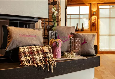 A stylish living room with decorative pillows and a warm blanket. In the background, large windows and a cozy atmosphere can be seen.