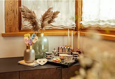 A stylish table with decorative objects, including flowers in a vase, candles, and colorful masks. The warm wood and color tones create a cozy atmosphere.