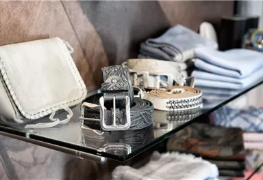 Eine elegante Auswahl an Accessoires auf einem Glasregal. Zu sehen sind eine Handtasche, mehrere Gürtel und gefaltete Tücher.