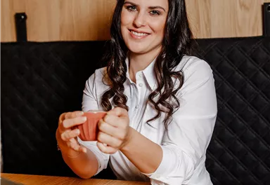 Eine Frau mit langen Haaren sitzt an einem Tisch und hält eine Tasse. Vor ihr liegt ein kleines Dessert auf einem Teller und ein Laptop steht auf dem Tisch.