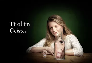 Eine Frau sitzt an einem Tisch, hinter einer Glasflasche mit einem Gesicht. Der Hintergrund ist dunkelgrün, und der Text zeigt "Tirol im Geiste".