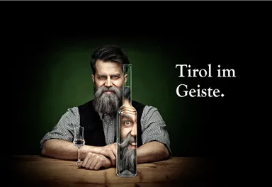 Ein Mann mit einem Bart hält einen langen Behälter, aus dem ein Gesicht erscheint. Neben ihm steht eine Flasche, und der Text sagt "Tirol im Geiste".