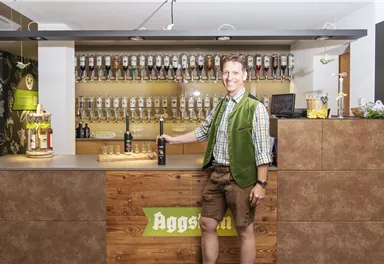 Ein freundlicher Mann im traditionellen bayerischen Outfit steht hinter der Bar. Hinter ihm sind zahlreiche Zapfhähne für Bier aufgereiht.