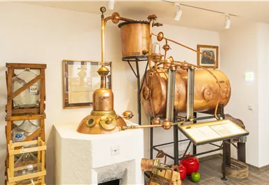 Eine alte Destillationsanlage mit Kupferkomponenten und verschiedenen Behältern. Im Hintergrund sind einige Informationsplakate zu sehen.