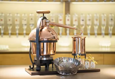 Eine kupferne Brennblase steht auf einem Tisch. Im Hintergrund sind mehrere Flaschen sichtbar.