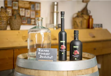 Eine Holztischplatte mit einer großen Glasflasche und zwei Flaschen Himbeerdestillat. Daneben steht ein Schild mit der Aufschrift "Reines Himbeer Destillat".