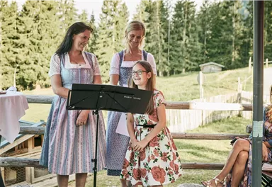 Three women in traditional attire stand on a terrace. A girl smiles and holds a paper for a performance.