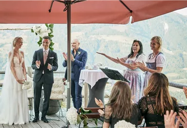 A wedding outdoors with a magical mountain landscape in the background. The bride and groom stand before guests and a ceremony, surrounded by flowers and a festive atmosphere.