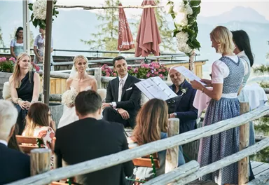 A beautiful outdoor wedding with mountains in the background. The bride and groom are seated surrounded by guests, while the officiant reads.