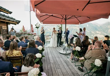 An outdoor wedding ceremony on a terrace with a view of the mountains. Guests are seated in chairs and applauding the couple.