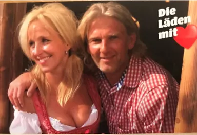 A smiling couple stands together in front of a wooden building. The woman is wearing a traditional Bavarian dirndl and the man is wearing a checkered shirt.