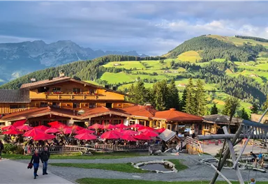 A cozy restaurant with a red sunshade in a picturesque mountain landscape. The surroundings are green and hilly, surrounded by mountains and a clear sky.