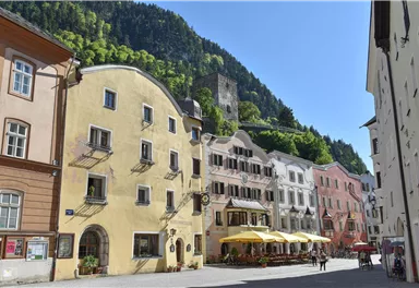 A picturesque square with colorful buildings and outdoor dining. In the background, there are green hills and a castle ruin.
