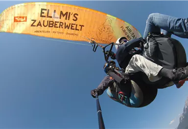 A paragliding flight over a picturesque landscape with blue skies. The paraglider carries the inscription "ELLMI'S MAGIC WORLD".