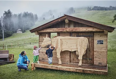 A wooden house with a wooden figure of a cow stands on a meadow. Two children and a woman stand in front of it and explore the figure.