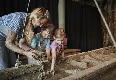 A woman shows two children how to work with clay. They are shaping something together in a workshop.