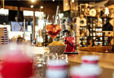 A stylish bar with various drinks on the counter. In the foreground, there is a glass with a colorful cocktail and a bowl of ice.