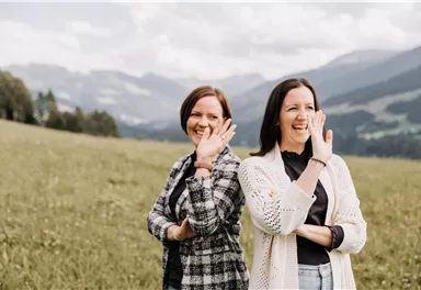 Zwei Frauen stehen in einer Wiese und winken lächelnd in die Kamera. Im Hintergrund sind grüne Hügel und Berge zu sehen.