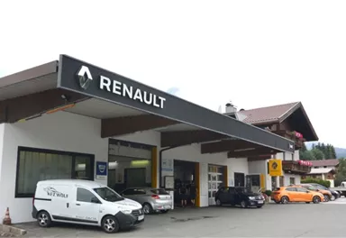 Eine Renault-Werkstatt mit mehreren Autos und einem großen Schild. Im Hintergrund sind Berge und ein traditionelles Gebäude zu sehen.