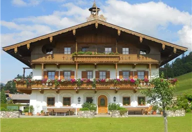 Ein traditionelles bayerisches Haus mit einer blühenden Balkonvegetation. Umgeben von grünen Wiesen und einem freundlichen Himmel.