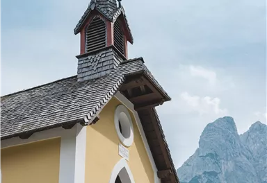 Eine kleine Kirche mit einem spitzen Turm und grauem Dach steht vor einer beeindruckenden Berglandschaft. Der Himmel ist leicht bewölkt und verleiht der Szene eine ruhige Atmosphäre.