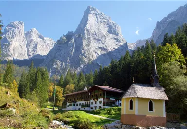 Eine malerische Berglandschaft mit hohen Gipfeln und üppigen Wäldern. Im Vordergrund steht eine kleine Kapelle und ein charmantes Holzgebäude.