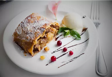 A delicious piece of apple strudel with powdered sugar, served on a white plate. Accompanied by a scoop of ice cream and fresh berries.