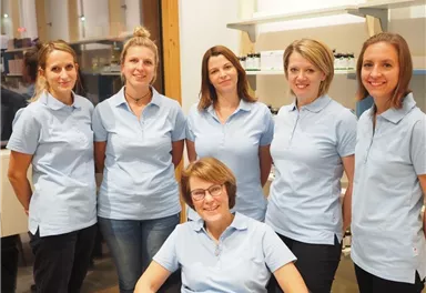 Eine Gruppe von sechs Personen in hellblauen Hemden steht zusammen in einem Raum. Eine Person sitzt im Vordergrund und lächelt freundlich.