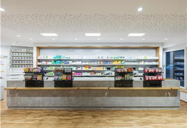 A modernly designed pharmacy interior with shelves full of medications. The concrete reception desk is in the foreground, providing a friendly atmosphere.