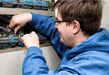 A person is working on an electrical switchgear. The man is wearing a blue sweater and looks focused.
