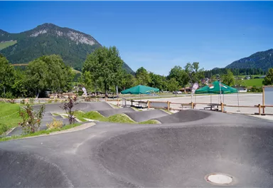 A modern skate park with curving ramps and a beautiful mountain landscape in the background. The surroundings are green and inviting, ideal for outdoor activities.