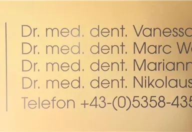 A golden background with the lettering of a dental practice. The names and titles of four dentists as well as a phone number are visible.