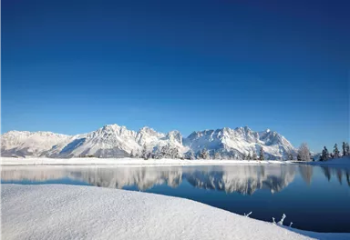 A winter landscape with snow-covered mountains and a clear blue sky. The calm lake reflects the impressive mountains.