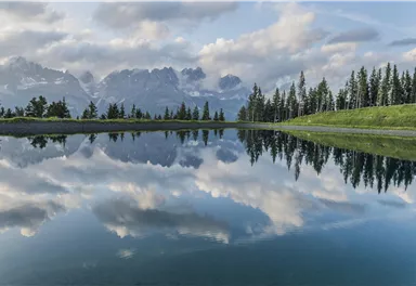 A calm lake reflects the clouds and mountains. Surrounded by trees, the landscape offers a peaceful atmosphere.