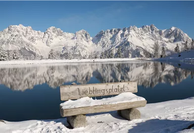 A snowy landscape with tall mountains in the background and a calm, reflective lake. A wooden bench with the inscription "Favorite Place" stands at the shore.