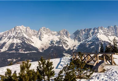 An impressive mountain landscape with snow-covered peaks and a clear blue sky. In the foreground, there is a wooden railing that provides a beautiful view of the surroundings.