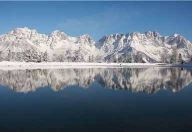 An impressive mountain landscape with snow-capped peaks and a tranquil lake in the foreground. The clear blue sky reflects in the water.