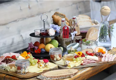 A lovingly arranged table with various types of cheese, sausage, and fresh fruit. There is also bread, jam, and pretty flowers as decoration.