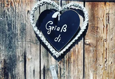 A heart-shaped sign hangs on a wooden wall and bears the inscription "Griaß di". The frame of the sign is made of woven material.