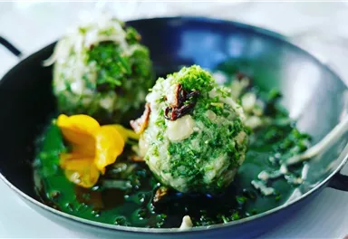 Two green dumplings on a black plate, surrounded by fresh herbs. An edible flower adds a splash of color.