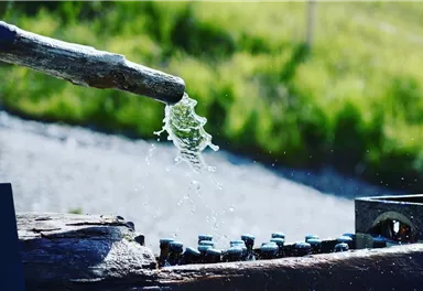 A water stream bubbles out of a wooden pipe. In the background, green meadows can be seen.